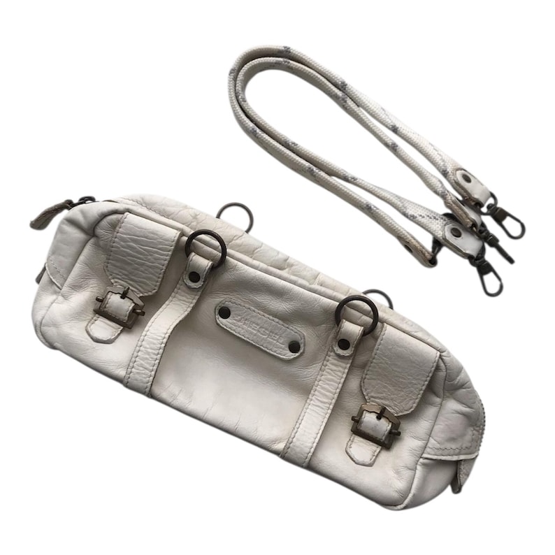 K&ouml;nnte beinhalten: Cremefarbene Lederhandtasche mit abnehmbarem Schulterriemen. Die Tasche hat zwei Vordertaschen mit Schnallenverschl&uuml;ssen, Metallringen und einen Rei&szlig;verschluss. Der Schulterriemen ist wei&szlig; mit einem grauen Muster.