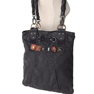 May include: Black denim tote bag with braided straps and metal hardware. The bag features the word "DIESEL" across the front in a mix of leather and metal letters. The bag has a structured design with a top handle and shoulder straps.
