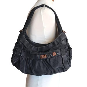 May include: A black hobo-style shoulder bag crafted from a textured fabric with a gathered, slouchy silhouette. The front features a horizontal strap adorned with metallic square hardware spelling out the brand name DIESEL in individual letters.