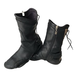 May include: A pair of black leather boots with a unique design. The boots have a zipper closure on the side, a strap with a buckle, and laces at the top. The soles are flat and flexible, and the boots appear to be unworn.