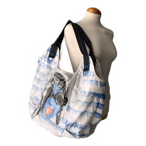 May include: A large white and light blue striped tote bag featuring a graphic print of a person wearing goggles and a helmet with a bright orange accent. The bag has two black shoulder straps with white stitching and the Diesel brand name printed.