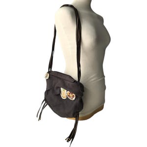 May include: A dark gray crossbody bag with a flap closure and long, dark brown straps. The bag features decorative patches of food items and gold-toned hardware. The bag is displayed on a mannequin.