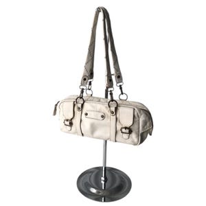 K&ouml;nnte beinhalten: Cremefarbene Handtasche mit zylindrischer Form, zwei kurzen Griffen und einem langen Schulterriemen. Die Tasche hat mehrere F&auml;cher, Metallbeschl&auml;ge und Schnallenverschl&uuml;sse. Die Tasche wird auf einem silbernen St&auml;nder pr&auml;sentiert.