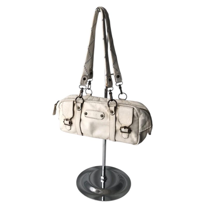 K&ouml;nnte beinhalten: Cremefarbene Handtasche mit zylindrischer Form, zwei kurzen Griffen und einem langen Schulterriemen. Die Tasche hat mehrere F&auml;cher, Metallbeschl&auml;ge und Schnallenverschl&uuml;sse. Die Tasche wird auf einem silbernen St&auml;nder pr&auml;sentiert.