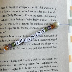 May include: A beaded bookmark with the word "INFINITY" spelled out in white letter beads. The bookmark has a pearl bead and colorful accent beads. The bookmark is resting on the pages of a book.