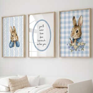 May include: Three framed prints featuring illustrations of a bunny rabbit. One print has the text "you'll never know dear how much we love you". The other two prints have a bunny rabbit wearing a blue jacket. The background of the prints is blue and white.