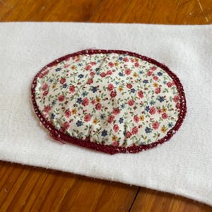 Op de afbeelding: Een ovaalvormige stoffen patch met een bloemenpatroon van rode, blauwe en gele bloemen op een witte achtergrond. De patch is afgezet met een donkerrode gehaakte rand en ligt op een witte stoffen ondergrond.