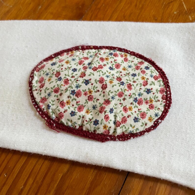 Op de afbeelding: Een ovaalvormige stoffen patch met een bloemenpatroon van rode, blauwe en gele bloemen op een witte achtergrond. De patch is afgezet met een donkerrode gehaakte rand en ligt op een witte stoffen ondergrond.