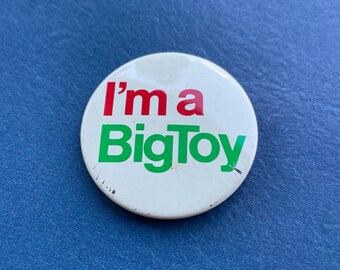 Op de afbeelding: Een ronde, witte button met de tekst "I'm a BigToy" in rode en groene letters. De button heeft een speld op de achterkant en staat tegen een blauwe achtergrond.