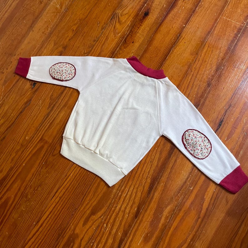 Op de afbeelding: Een cr&egrave;mekleurige sweatshirt met lange mouwen en bordeauxrode accenten. De manchetten en kraag zijn bordeauxrood en bloemige patches sieren de ellebogen. De sweatshirt is gemaakt van zacht materiaal en is geschikt voor kinderen.