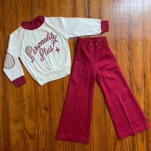 May include: A vintage cream-colored outfit with a long-sleeved top and flared pants. The top has burgundy trim, a floral elbow patch, and the words "Personality Plus" in burgundy script. The pants are a matching burgundy color.