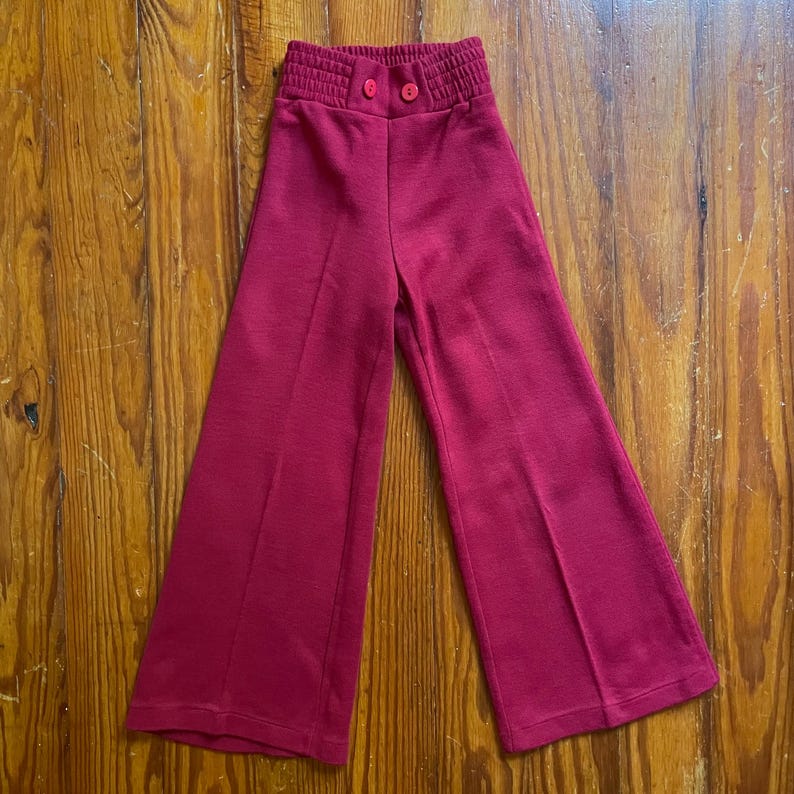 Op de afbeelding: Een paar magenta uitlopende broeken. De broek heeft een hoge, elastische tailleband met twee bijpassende knopen. De stof lijkt een zachte tricot te zijn en de pijpen zijn wijd en vloeiend.