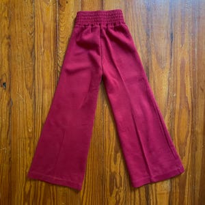 Op de afbeelding: Een rode broek met wijde pijpen en een gerimpelde tailleband. De broek is gemaakt van een zacht materiaal en heeft een wijd beenontwerp. De broek wordt gepresenteerd op een houten oppervlak.