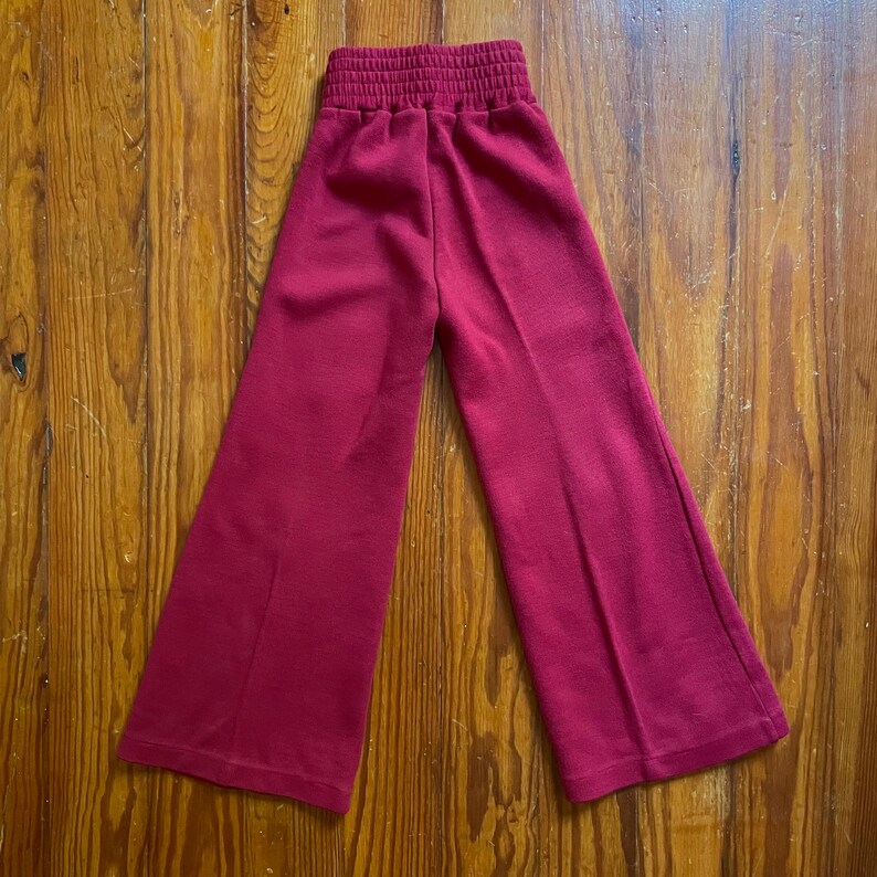 Op de afbeelding: Een rode broek met wijde pijpen en een gerimpelde tailleband. De broek is gemaakt van een zacht materiaal en heeft een wijd beenontwerp. De broek wordt gepresenteerd op een houten oppervlak.
