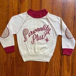 Op de afbeelding: Cr&egrave;mekleurige sweatshirt met een bordeauxrode col, manchetten en bloemen elleboogstukken. De woorden "Personality Plus!" zijn in bordeauxrood script op de voorkant geborduurd. De sweatshirt heeft een vintage esthetiek.