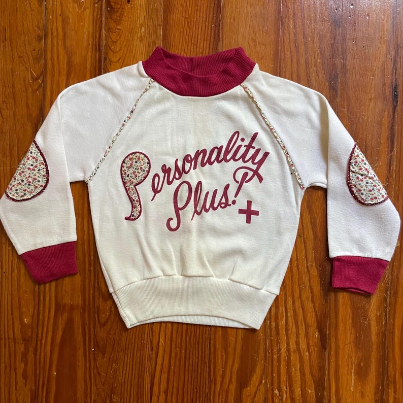 Op de afbeelding: Cr&egrave;mekleurige sweatshirt met een bordeauxrode col, manchetten en bloemen elleboogstukken. De woorden "Personality Plus!" zijn in bordeauxrood script op de voorkant geborduurd. De sweatshirt heeft een vintage esthetiek.