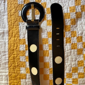 May include: A black patent leather belt featuring a large circular buckle with a gold-toned metal prong. The belt strap is adorned with evenly spaced white circular cutouts and includes a series of rectangular adjustment holes at the end.