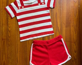 Op de afbeelding: Een rood-wit gestreept T-shirt met witte knopen en een bijpassende rode short met witte bies. De outfit is gemaakt van een zacht materiaal en ligt op een houten oppervlak.