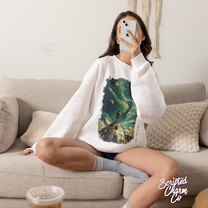May include: A white crewneck sweatshirt featuring a graphic print of a lone figure standing on a stone tower looking up at a swirling green and blue nebula with stars and planets. The text "SAVE THE STARS" is printed vertically along the side.