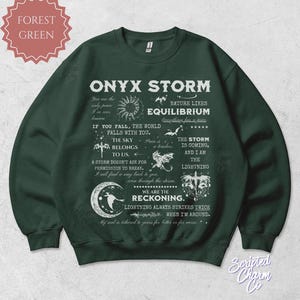 May include: Forest green sweatshirt with the text "ONYX STORM" and various quotes and illustrations. The design includes a sun, moon, stars, and other celestial elements. The sweatshirt is a crew neck style.