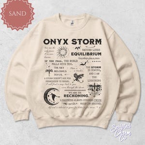 May include: Sand-colored sweatshirt with the text "ONYX STORM" and various graphics, including a sun, stars, and a bat. The design features several lines of text with inspirational quotes. The sweatshirt is a crew neck.