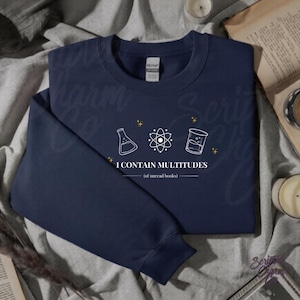 May include: Navy blue sweatshirt with white science-themed graphics and the text "I CONTAIN MULTITUDES (of unread books)". The design includes a flask, an atom, and a beaker. The sweatshirt is folded on a soft surface.