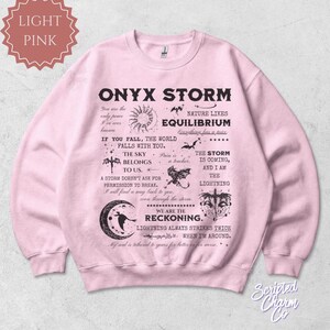 May include: Light pink sweatshirt with the text "ONYX STORM" and various graphics. The design includes phrases like "The sky belongs to us" and "We are the reckoning."