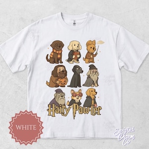 Puede incluir: Camiseta blanca con una ilustración de dibujos animados de perros disfrazados de personajes de una serie de fantasía. La frase "Hairy Paw-ter" está impresa en oro debajo de los perros. Una etiqueta roja y blanca dice "WHITE".