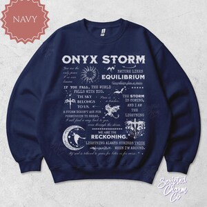 May include: Navy blue sweatshirt with the text "ONYX STORM" and various celestial and nature-themed graphics. The design includes phrases like "The sky belongs to us" and "We are the reckoning."