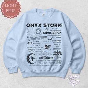 May include: Light blue Onyx Storm sweatshirt with black text and graphics. The design includes phrases like "The Sky Belongs to Us" and "We Are The Reckoning." Features a crew neck and long sleeves. The sweatshirt is a comfortable, casual wear item.