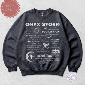 May include: Dark heather gray sweatshirt with the text "ONYX STORM" and other phrases and graphics in white. The sweatshirt has a crew neck and long sleeves. The brand name "Scripted Charm Co" is in the bottom right corner.
