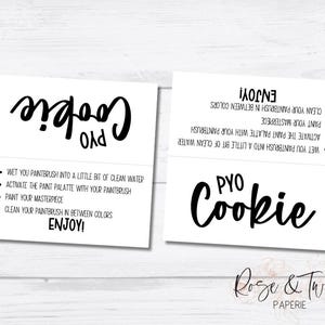 May include: Two white cards with black text. The top card says "PYO Cookie!" and the bottom card has instructions for painting a cookie. The instructions are: "Wet you paintbrush into a little bit of clean water. Activate the paint palette with your paintbrush. Paint your masterpiece. Clean your paintbrush in between colours. Enjoy!"