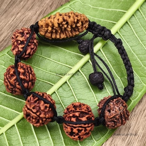 May include: A bracelet featuring six brown, textured, spherical and oval rudraksha beads strung on a black braided cord. The cord includes a sliding knot mechanism with two smaller black beads at the ends. The text RamratanShop is visible.
