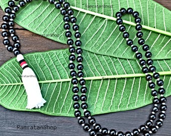 May include: Black beaded necklace with a white tassel and red and black accents. The necklace is made of small, round, black beads strung together with white string. The tassel is white and fluffy. The necklace is displayed on a green leaf.