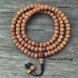 Puede incluir: Un collar mala hecho de cuentas marrones texturizadas. Las cuentas están ensartadas en tres hilos, formando una forma circular. Una cuenta tallada más grande y una borla están en la parte inferior. El texto "RamratanShop" es visible.