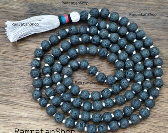 May include: A dark gray lava stone mala bead necklace with a white tassel. The beads are strung on white cord. The tassel has a white base with a band of red, blue, and black. The necklace is displayed on a wooden surface.