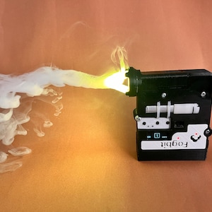 Puede incluir: Un dispositivo en blanco y negro con la palabra "Fogbit", que emite una corriente de humo blanco con un brillo amarillo. El dispositivo tiene forma rectangular con varios botones y detalles.