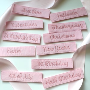 May include: Pink fabric tags embroidered with various celebratory phrases, including "Just Born," "Valentines," and "Halloween." A light pink ribbon weaves through the tags, creating a decorative arrangement. The tags are ideal for gift wrapping or craft projects.