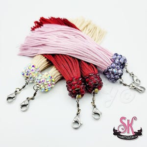 May include: A collection of five decorative tassels featuring long, thin fabric strands in shades of light pink, red, beige, and cream. Each tassel is topped with a spherical bead encrusted with sparkling crystals and includes a silver metal lobster clasp.