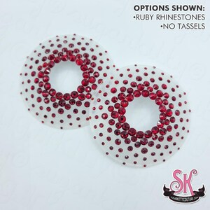 May include: Two circular, translucent pasties decorated with ruby red rhinestones in a gradient pattern. Text in the image reads: OPTIONS SHOWN: RUBY RHINESTONES, NO TASSELS. The logo SK is visible in the bottom right corner.