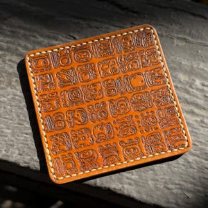 May include: Square, tan leather coaster with a detailed embossed pattern of ancient glyphs. The edges are stitched with a light-colored thread. The coaster is designed to protect surfaces from drink condensation and heat.