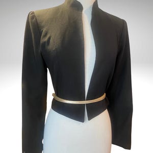 Peut inclure: Un blazer noir avec un col montant et des manches longues. Une ceinture dorée cintre la taille. La veste est présentée sur un mannequin blanc, mettant en valeur sa coupe ajustée et son design.