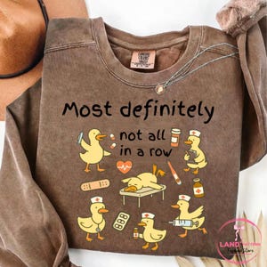 Pu&ograve; includere: Felpa marrone a maniche lunghe con la scritta "Most definitely not all in a row". Il design presenta anatre dei cartoni animati con cappelli da infermiera e forniture mediche. Una collana e un cappello sono visibili.
