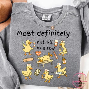 Pu&ograve; includere: Una felpa grigia con la scritta "Most definitely not all in a row" e anatre dei cartoni animati con cappelli da infermiera. Le anatre sono raffigurate con forniture mediche, tra cui siringhe, bende e flaconi di medicinali. Marchio Comfort Colors.