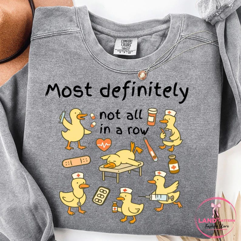 Pu&ograve; includere: Una felpa grigia con la scritta "Most definitely not all in a row" e anatre dei cartoni animati con cappelli da infermiera. Le anatre sono raffigurate con forniture mediche, tra cui siringhe, bende e flaconi di medicinali. Marchio Comfort Colors.