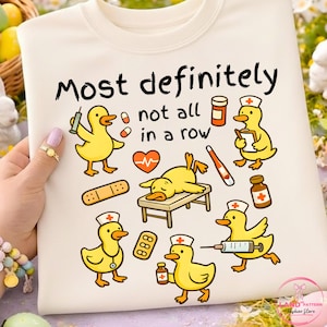 Peut inclure: Sweat-shirt crème avec un motif de canards jaunes de dessin animé dans des rôles médicaux, entourés de fournitures médicales. Le texte "Most definitely not all in a row" est imprimé au-dessus des canards.