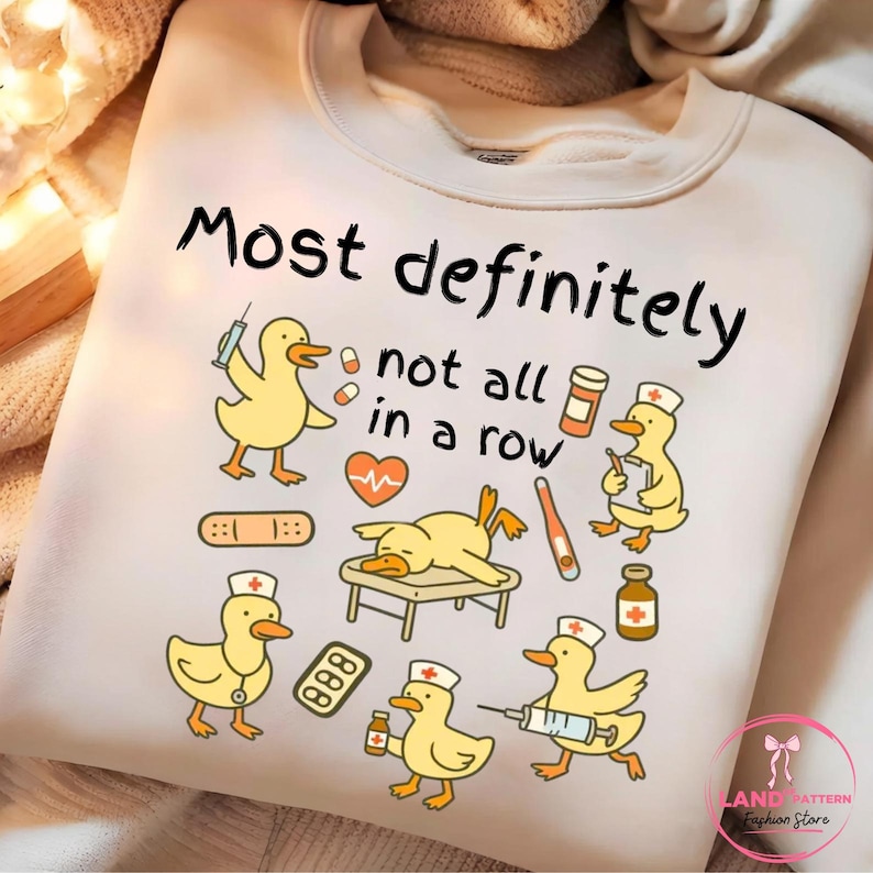 Pu&ograve; includere: Felpa color crema con una grafica giocosa di anatre gialle vestite da professionisti medici. Il design include siringhe, pillole e la scritta "Most definitely not all in a row".