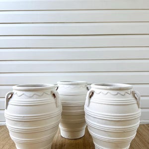 May include: Three off-white ceramic vases with a textured, ribbed design and small handles. The vases have a classic shape and a decorative wavy pattern near the top. They are arranged on a woven mat against a white background.