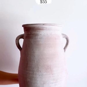 May include: A large, raw-finish Zafora Urn in a light pink hue, with two curved handles and a wide opening. The urn is held by a person. The text "Zafora Urn raw finish" is visible.