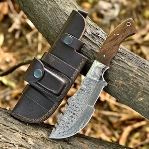 May include: A Damascus steel knife with a wooden handle and a brown leather sheath. The knife blade has a unique pattern and a serrated edge. The sheath has a snap closure and belt loop. The knife and sheath are resting on a tree branch.